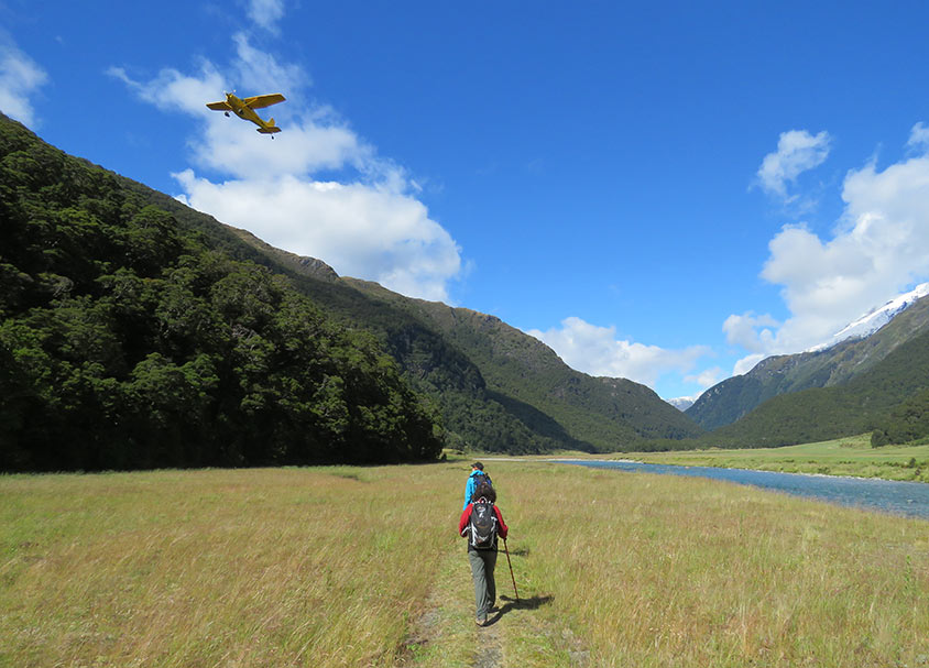 Two hikers on the trail, a river running beside, and a small aircraft flying overhead.