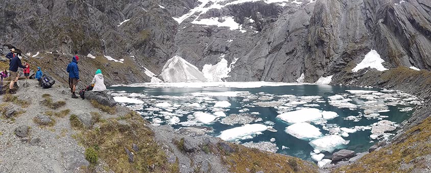 Panoramic shot of glacial lake, icebergs floating, and a group sitting on the shore.